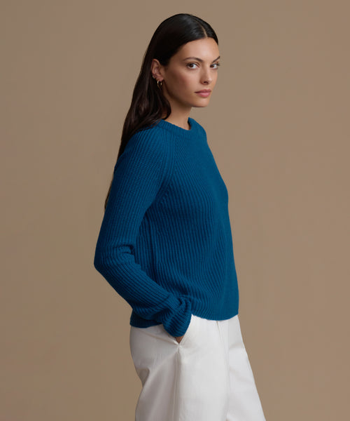 Woman wearing a blue cashmere sweater and white pants against a brown background