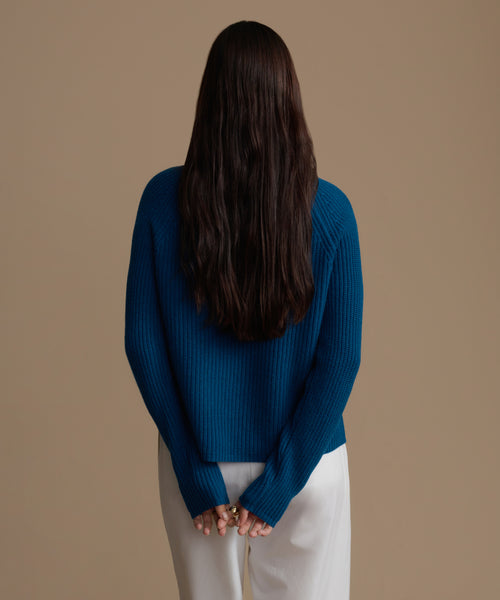 Woman wearing a blue cashmere sweater and white pants against a brown background