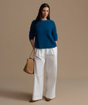 Woman wearing a blue cashmere sweater and white pants, holding a brown bag on a beige background