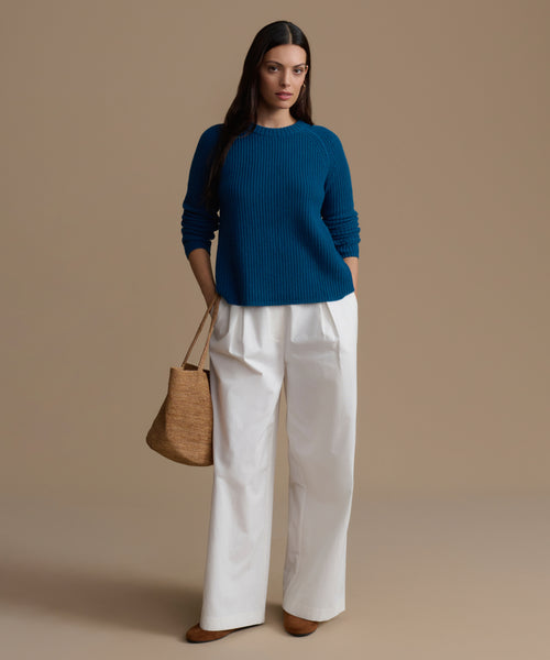 Woman wearing a blue cashmere sweater and white pants, holding a brown bag on a beige background
