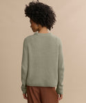 A person with short curly hair stands with their back to the camera against a beige background, wearing the Cashmere Fisherman Sweater in sage and brown pants.
