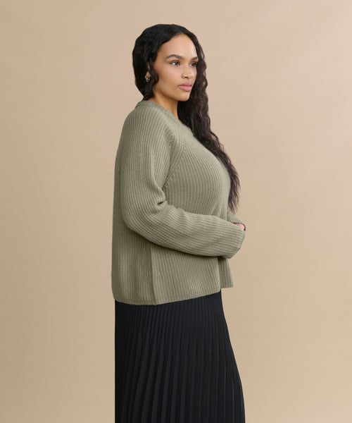 A woman with long, wavy dark hair stands in profile against a beige background, wearing the Cashmere Fisherman Sweater in sage and a long black pleated skirt. She appears relaxed, with her hands resting gently in front of her.