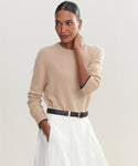 A woman with dark hair pulled back wears the Flynn Cashmere Crewneck and a white skirt with a black belt, standing with one hand in her pocket and looking to the side against a plain background.
