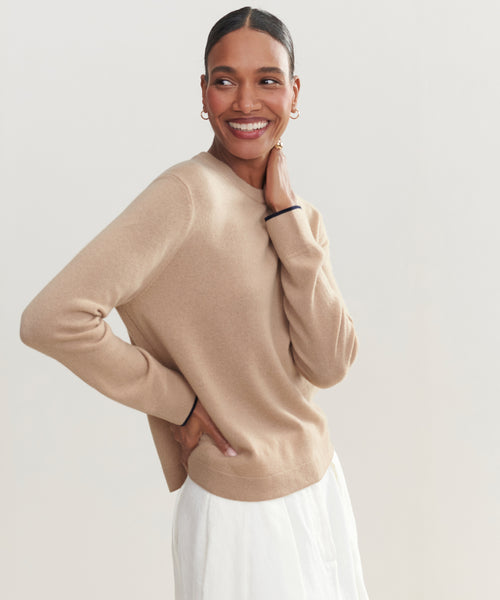 A woman smiles with one hand on her hip, wearing the Flynn Cashmere Crewneck in beige and a white skirt, accessorized with gold hoop earrings. The background is plain and light-colored.