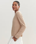 A woman with dark hair in a sleek bun wears the Flynn Cashmere Crewneck sweater in beige with a white skirt, standing sideways and smiling softly at the camera against a plain light background.