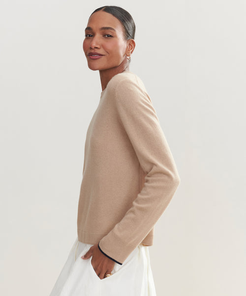 A woman with dark hair in a sleek bun wears the Flynn Cashmere Crewneck sweater in beige with a white skirt, standing sideways and smiling softly at the camera against a plain light background.