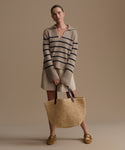Woman wearing a striped cashmere sweater and holding a woven bag against a beige background