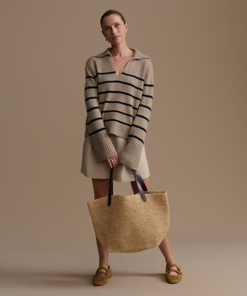 Woman wearing a striped cashmere sweater and holding a woven bag against a beige background