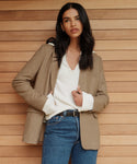 A woman with long dark hair stands before a wooden wall, wearing the Cashmere Frances Polo in ivory beneath a tan blazer and relaxed blue jeans, her hands tucked in her pockets and her expression neutral.