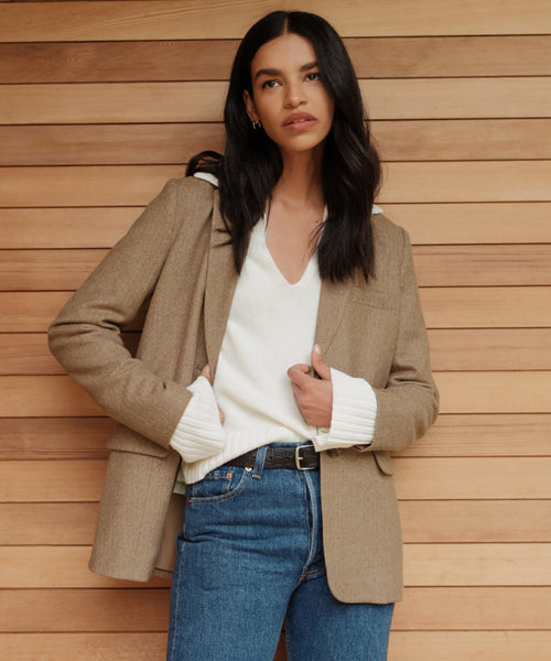 A woman with long dark hair stands before a wooden wall, wearing the Cashmere Frances Polo in ivory beneath a tan blazer and relaxed blue jeans, her hands tucked in her pockets and her expression neutral.
