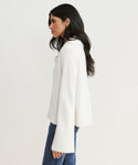 A woman with long dark hair stands in profile, wearing the Cashmere Frances Polo—a relaxed fit ivory knit sweater with wide ribbed cuffs—paired with blue jeans against a plain light background.