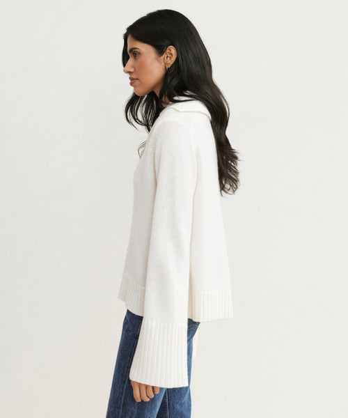 A woman with long dark hair stands in profile, wearing the Cashmere Frances Polo—a relaxed fit ivory knit sweater with wide ribbed cuffs—paired with blue jeans against a plain light background.