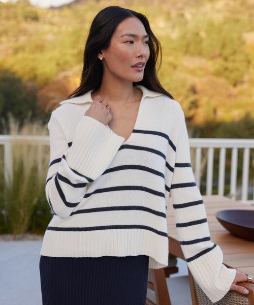 A woman with long dark hair stands outdoors, wearing the Cashmere Frances Polo—a relaxed-fit white knit sweater with black and navy stripes—and a navy skirt. She touches her collar, gazing to the side by a railing with trees behind her.