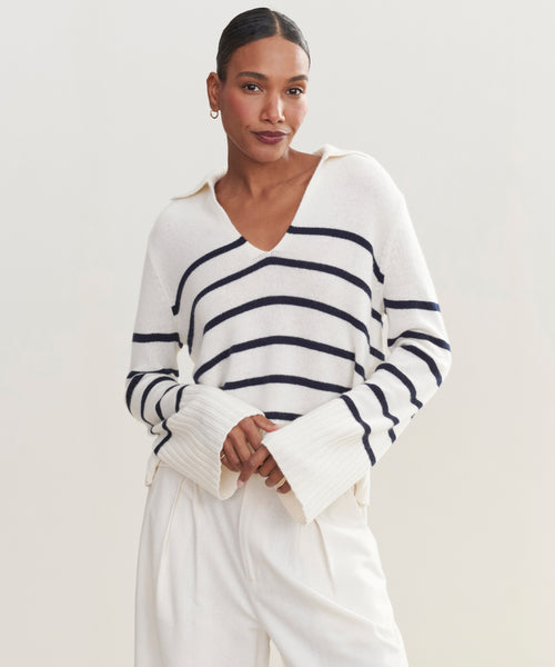 A woman wears the Cashmere Frances Polo—a relaxed fit knit white sweater with bold navy stripes and wide sleeves—paired with white pants. She stands against a plain light background, hair pulled back, looking confidently at the camera.
