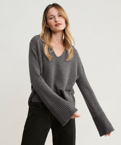 Woman with long blonde hair wears the Cashmere Frances Polo in dark gray, featuring a relaxed fit and wide ribbed cuffs, paired with black pants against a plain light background.