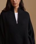 A close-up of a woman wearing a black ribbed knit half-zip sweater with a stand collar, layered over a white top, against a solid tan background.