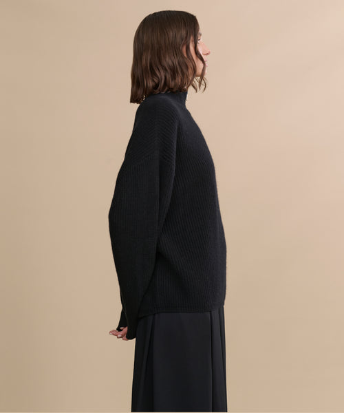 A woman with shoulder-length brown hair stands in profile against a plain beige background, wearing a relaxed Cashmere Half Zip sweater and a long black skirt.