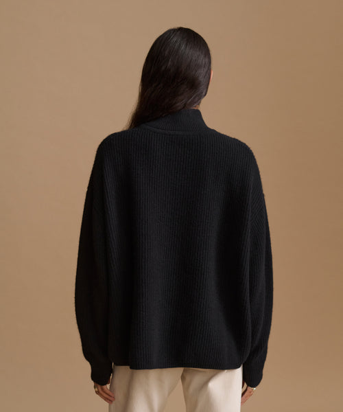 A woman viewed from the back wearing a black ribbed knit mock-neck sweater and cream pants, set against a solid warm tan studio background.