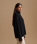 A woman is shown in profile wearing a black ribbed knit mock-neck sweater and cream-colored pants, set against a solid warm beige studio background.