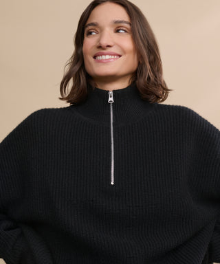 A woman with shoulder-length brown hair smiles while wearing the Cashmere Half Zip, a relaxed high-collar sweater with a zip-up front, set against a plain beige background.
