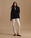A woman modeling a black long-sleeve zip-front sweater over a white top, paired with cream-colored trousers and black flats, against a solid beige studio background.