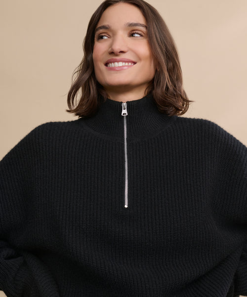 A woman with shoulder-length brown hair smiles while wearing the Cashmere Half Zip, a relaxed high-collar sweater with a zip-up front, set against a plain beige background.