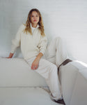 A woman wearing a cream-colored ribbed knit half-zip sweater and matching wide-leg sweatpants, sitting on a white sofa against a white brick wall.