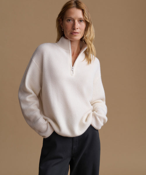 A woman wearing a cream-colored ribbed knit quarter-zip sweater and dark trousers, standing against a solid beige background.
