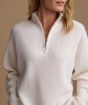 A close-up of a woman wearing a cream-colored ribbed knit quarter-zip sweater against a warm beige background.