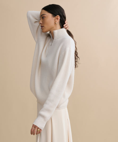 A woman with long dark hair in loose waves models the Cashmere Half Zip sweater and matching skirt, standing sideways against a beige background, one hand resting on the back of her head.