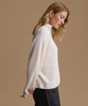 A woman wearing a ribbed knit mock neck sweater in off-white, paired with dark trousers, posed against a solid warm beige background.