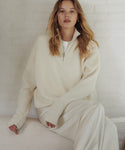 A woman with blonde hair sits on a light-colored sofa wearing a cream ribbed knit half-zip sweater and matching wide-leg trousers over a white top.