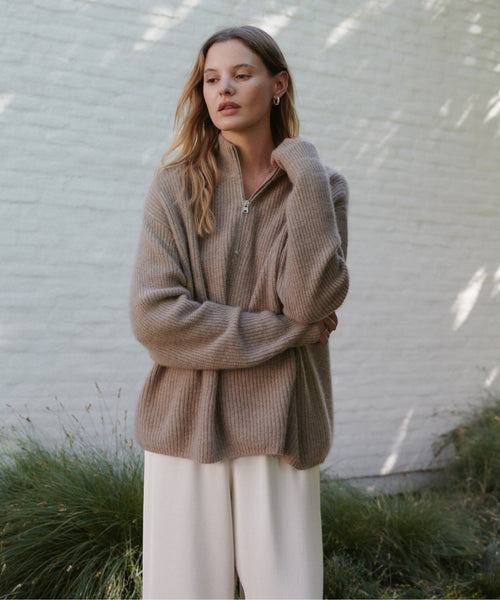 A woman wearing a ribbed knit half-zip sweater in taupe and white wide-leg pants, standing outdoors against a white brick wall with soft shadows.