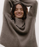A woman with brown hair smiles with her eyes closed, arms raised, wearing the cozy, oversized Cashmere Jackie Sweater against a plain light background.