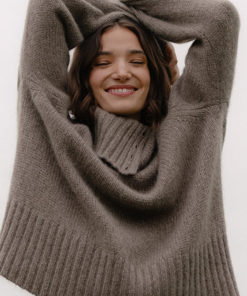 A woman with brown hair smiles with her eyes closed, arms raised, wearing the cozy, oversized Cashmere Jackie Sweater against a plain light background.