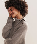 A person with curly hair smiles with their eyes closed, covering part of their face. Theyre wearing the Cashmere Jackie Sweater in light gray, a luxe knit featuring a high collar and distinctive button-shoulder detailing.