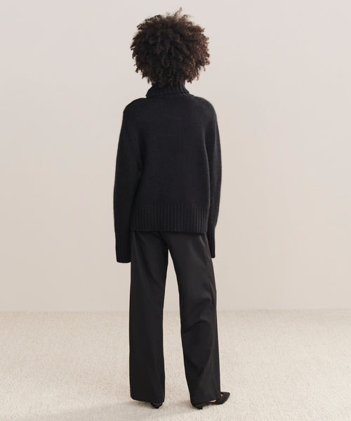 A person with curly hair, seen from behind, is wearing the Cashmere Jackie Sweater in black with loose black pants, standing on a light-colored carpet against a plain neutral wall.