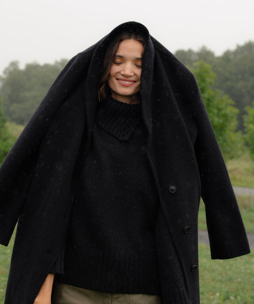 A woman stands outdoors on a cloudy day, smiling with her eyes closed while holding a black coat over her head. She wears the luxury Cashmere Jackie Sweater and green pants; trees fill the background.