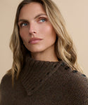 A woman with wavy blonde hair gazes upward thoughtfully, wearing the Cashmere Jackie Sweater in brown with button-shoulder detailing, set against a plain beige background.