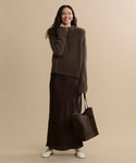A woman smiles against a beige background, wearing the Cashmere Jackie Sweater in brown with button-shoulder detail, a long dark brown skirt, white shoes, and carrying a matching brown tote bag.