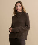 A woman with wavy, shoulder-length hair wears the Cashmere Jackie Sweater—a luxe brown knit with button-shoulder detailing—and a dark skirt, standing against a beige background and looking directly at the camera with a neutral expression.
