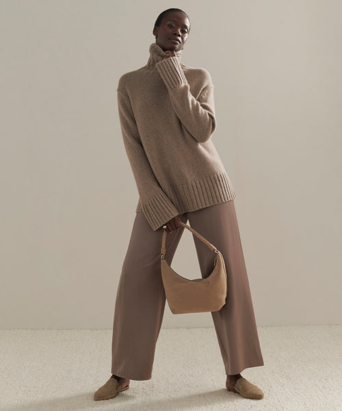 Wearing the Cashmere Jet Turtleneck, wide-legged mauve pants, and beige shoes, a person stands holding a matching beige handbag against a neutral background.