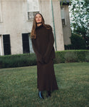 A woman with long brown hair stands on grass before a beige house, wearing the Cashmere Jet Turtleneck, a matching pleated skirt, and black boots. She looks calmly at the camera with her hands clasped.