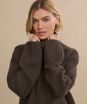 A woman with blonde hair wears the Cashmere Jet Turtleneck, holding the collar with both hands. She looks at the camera with a neutral expression, accessorized with gold rings and small hoop earrings against a beige background.