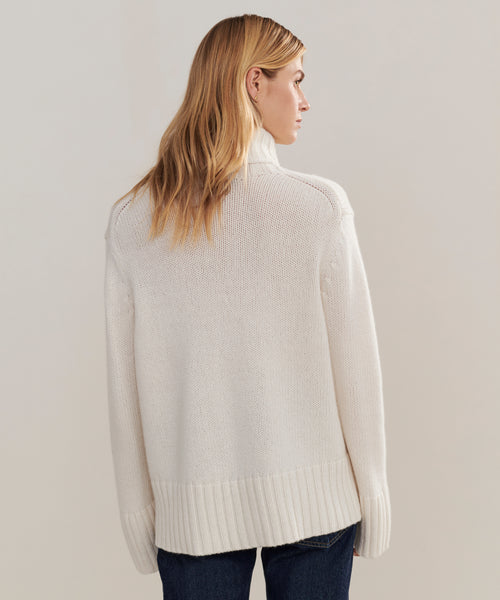 A person with long, blonde hair stands with their back to the camera against a plain light background, wearing the oversized Cashmere Jet Turtleneck and dark jeans.