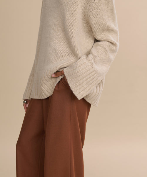 A person wearing the Cashmere Jet Turtleneck with long sleeves and brown wide-leg pants stands against a beige background, one hand tucked into their pocket.
