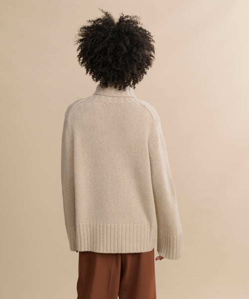 A person with curly hair is seen from behind, wearing the Cashmere Jet Turtleneck in beige and brown pants, standing against a plain light beige background.