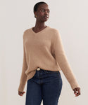 A woman in the Cashmere Kate V-Neck, a beige luxury sweater, and dark blue jeans stands against a plain light background, looking confidently to her left.