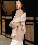 A woman with dark hair wears the Cashmere Kate V-Neck sweater draped over her shoulders and a cream top, standing outdoors in front of a wooden building and glancing back over her shoulder.
