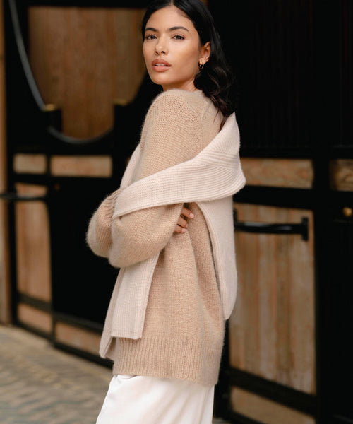 A woman with dark hair wears the Cashmere Kate V-Neck sweater draped over her shoulders and a cream top, standing outdoors in front of a wooden building and glancing back over her shoulder.
